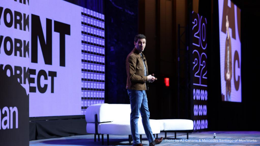 Male presenter on a stage in a brown jacket speaks to an audience, with a white couch on stage and tall screens projecting graphics in the background.