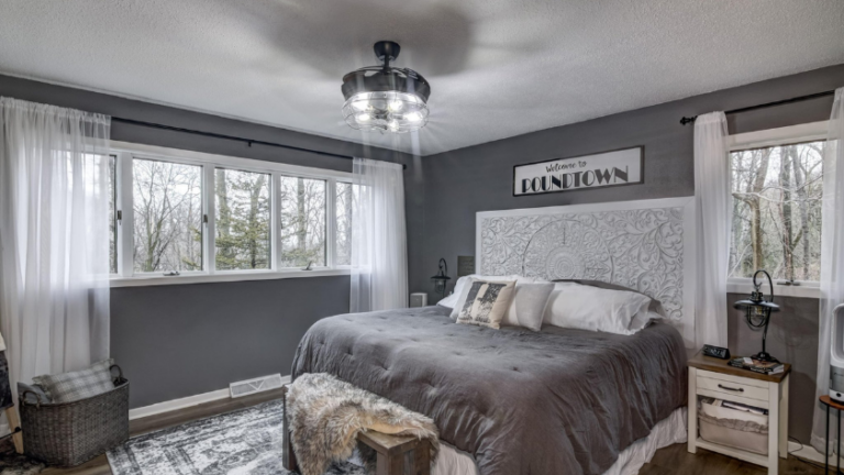 Cozy modern bedroom with dark gray walls, a large bed with an ornate white headboard, gray bedding, and a sign that reads “Welcome to Poundtown.”