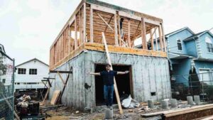 Man stands with arms outstretched in front of a two-story house under construction, with wooden framing atop a concrete foundation.