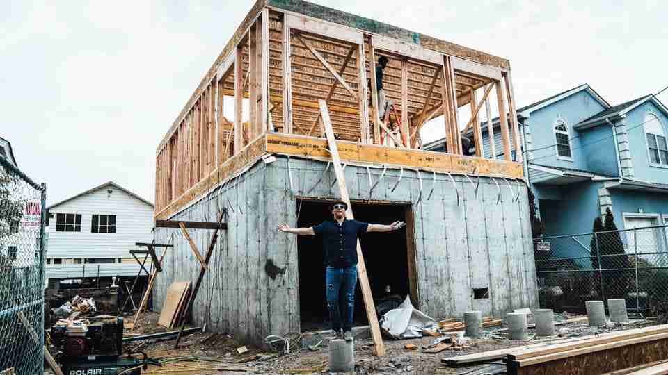 Man stands with arms outstretched in front of a two-story house under construction, with wooden framing atop a concrete foundation.