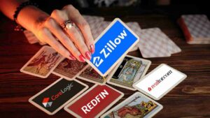 Hand with manicured nails and a ring places a blue Zillow card among scattered real estate brand cards on a wooden table surface.