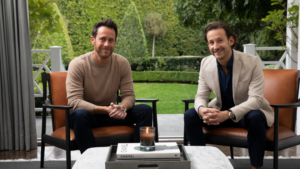 Two men sit on orange armchairs in a garden patio, smiling at the camera; a lit candle sits on the coffee table between them.