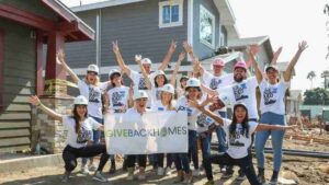 Group of volunteers in hard hats posing on a construction site, holding a 'Give Back Homes' banner and cheering.