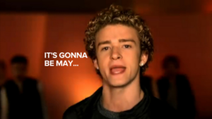 Close-up of a young man singing with the caption 'IT'S GONNA BE MAY...' on the left against a warm orange background.