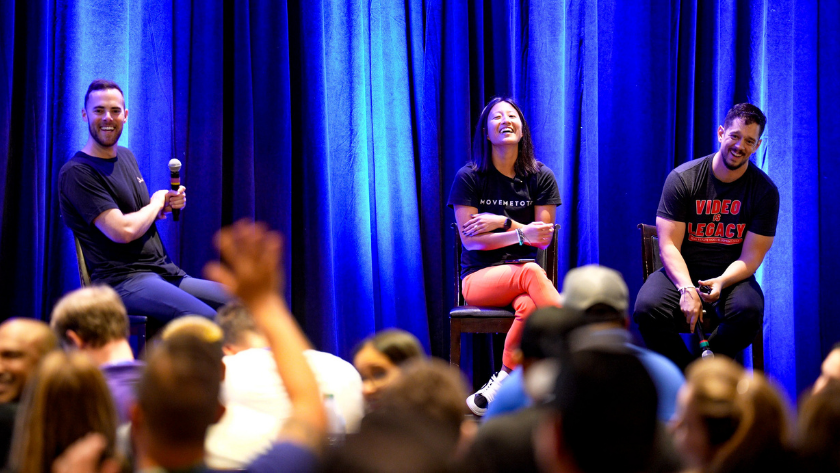 Three panelists on a blue-curtained stage; left man with a microphone, center woman laughing, right man smiling.
