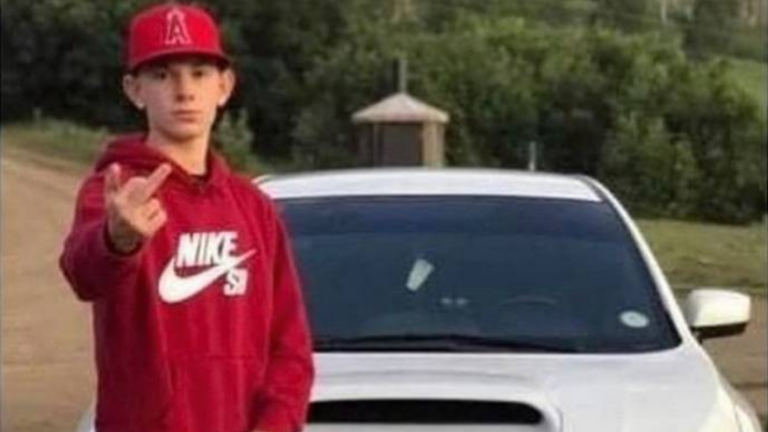 Teen in a red Nike hoodie and cap points at the camera while standing in front of a white car on a road.