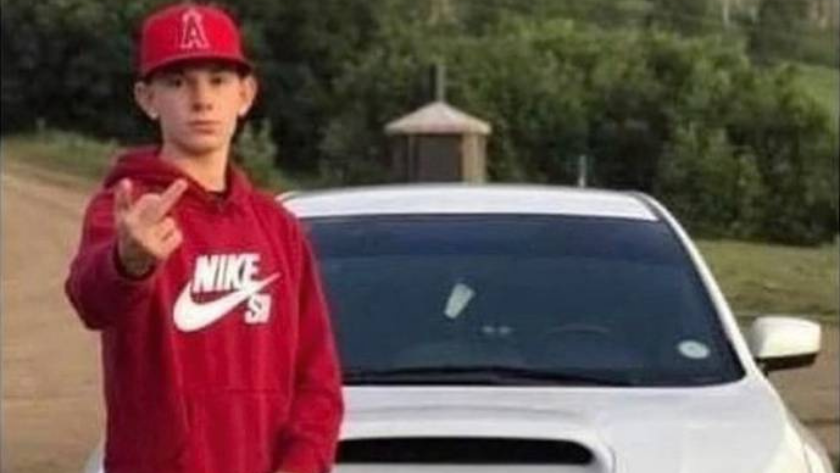 Teen in a red Nike hoodie and cap points at the camera while standing in front of a white car on a road.