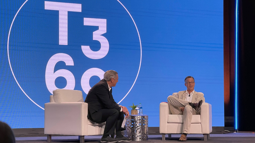 Two men sit in white armchairs on a stage during a talk show, with a large blue screen behind them displaying a circular logo and the numbers T3 60/0. Another man speaks while the other listens, a small table with water bottles and a plant between them.