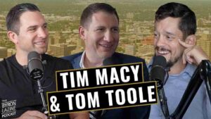 Three men in a podcast studio with microphones; bold banner reads Tim Macy & Tom Toole against a city backdrop.