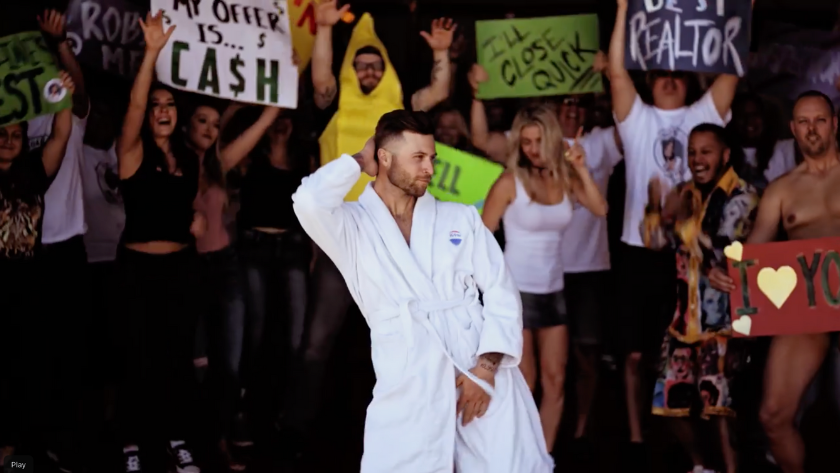 Man in a white robe poses with one hand behind his head in front of a cheering crowd holding colorful protest signs.
