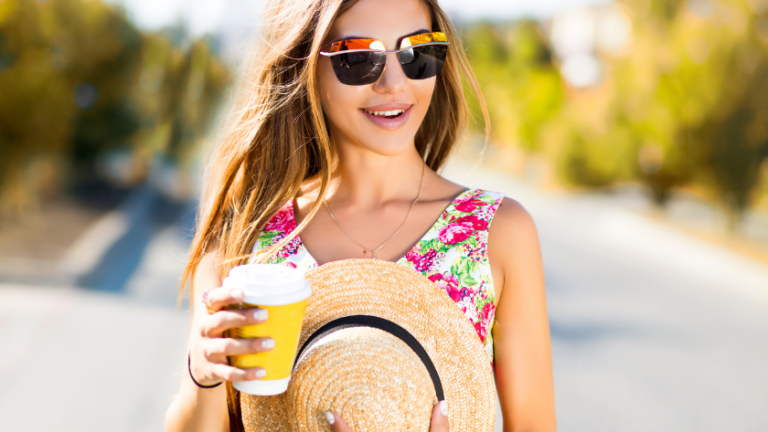 Smiling woman in a floral dress wearing sunglasses, holding a straw hat and a to-go coffee cup outdoors on a sunny day.