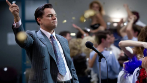 Man in a pinstriped suit raises a finger while speaking into a microphone as a cheering crowd tosses confetti in celebration behind him.