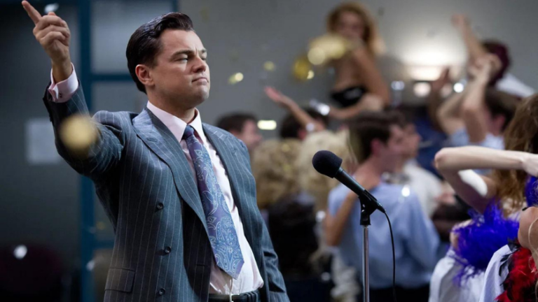 Man in a pinstriped suit raises a finger while speaking into a microphone as a cheering crowd tosses confetti in celebration behind him.