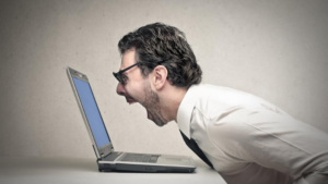 Man in a white shirt leans toward a laptop, shouting in frustration at the screen