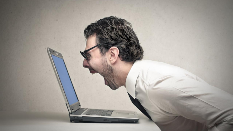 Man in a white shirt leans toward a laptop, shouting in frustration at the screen