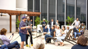 Group of people seated and standing at an outdoor meeting, listening to a speaker in a courtyard setting with chairs and plants.