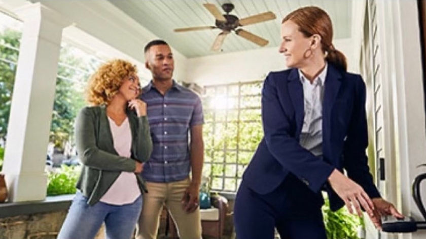 Three coworkers watch as a woman in a navy business suit demonstrates something near a doorway in a bright living room.