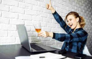 Woman at a desk with laptop, raising a thumbs-up and holding an orange drink, celebrating online success.