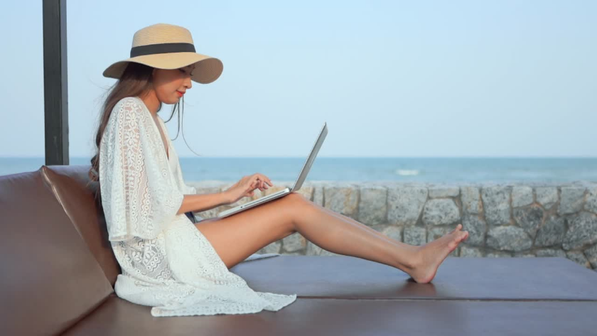 Woman in a white lace dress and wide-brim hat sits on an outdoor couch by the sea, typing on a laptop.