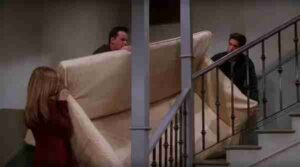 Three people carry a large sofa up a residential staircase, working together to move it upstairs.