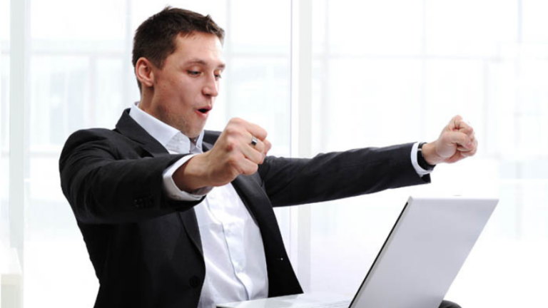 Businessman in a dark suit celebrating with fists raised in front of a laptop in a bright office