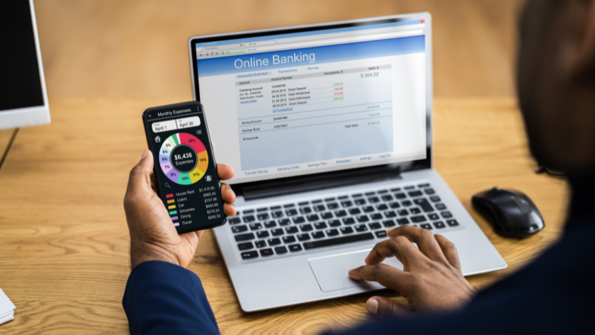 Person holds a smartphone displaying a colorful pie/donut chart beside a laptop showing an online banking page, on a wooden desk.