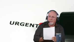 Host with headset microphone speaks into a paper sheet in a studio; a large 'URGENT' sign appears on the white background behind him.