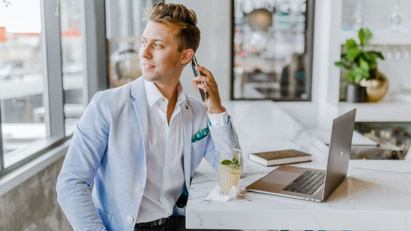 Man in a light blue blazer talks on his cellphone at a marble counter with a laptop nearby and an iced drink.