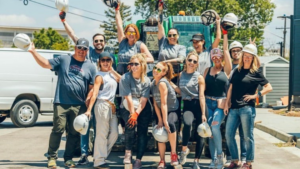 Team of utility workers posing for a group photo on a construction site, wearing hard hats and safety gear with a truck and equipment in the background.