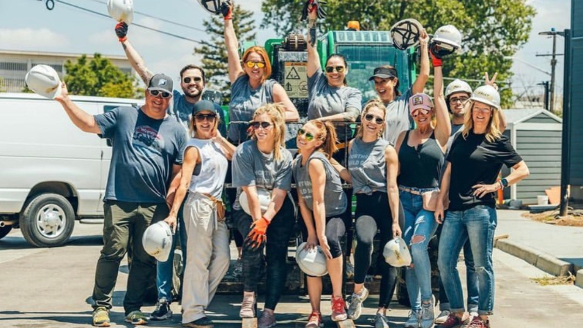 Team of utility workers posing for a group photo on a construction site, wearing hard hats and safety gear with a truck and equipment in the background.
