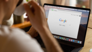 Person using a laptop at a wooden table with the Google homepage visible on the screen, holding a cup near their mouth.