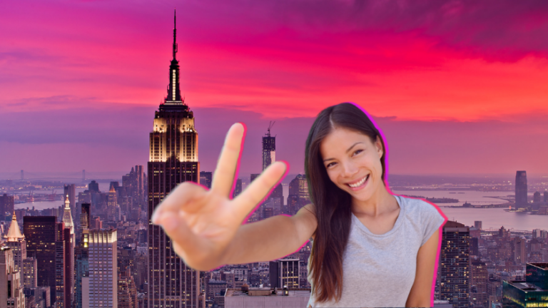 Smiling woman in gray t-shirt flashing a peace sign in front of a New York City skyline at sunset.