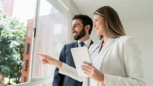 Two business professionals looking out a window; the woman points toward something outside while holding a tablet.