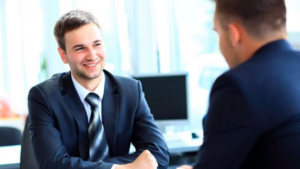 Two businessmen in suits shake hands across a desk, smiling during a meeting in a bright office.