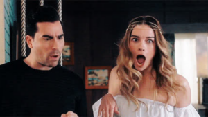 A man and a surprised woman stand indoors, both with wide eyes and open mouths.