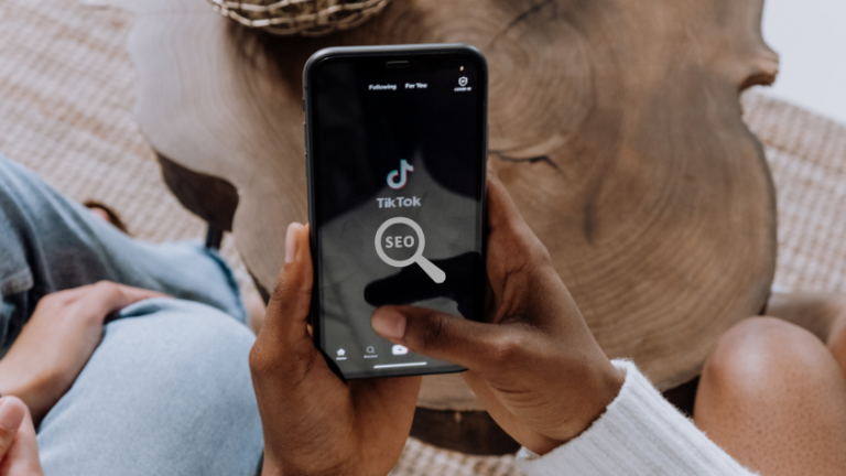 Person holding a smartphone displaying the TikTok app with a magnifying-glass icon labeled SEO in the foreground.