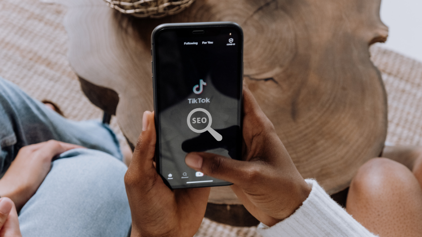 Person holding a smartphone displaying the TikTok app with a magnifying-glass icon labeled SEO in the foreground.