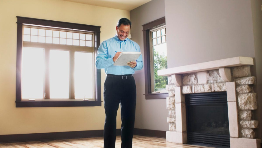 Man in a blue shirt with a clipboard inspecting a bright living room with a stone fireplace.