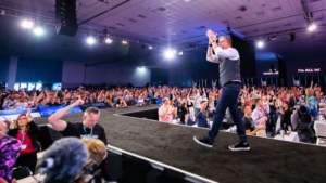 Male presenter walks along stage edge as crowd raises hands at a conference in a large indoor venue.
