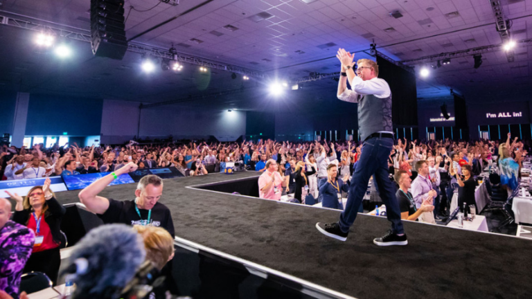 Male presenter walks along stage edge as crowd raises hands at a conference in a large indoor venue.