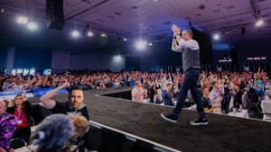 Speaker in a vest walks along the edge of a stage as a large audience cheers at a conference hall with bright lights overhead.