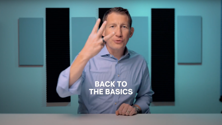 Presenter in a light blue shirt sits at a white table in a studio with teal walls and acoustic panels, gesturing with three fingers; overlay text reads 'Back to the Basics'