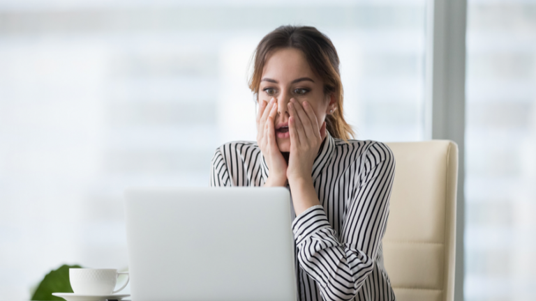 Woman in a striped blouse sits at a desk, covering her mouth while looking at a laptop.