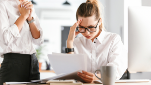 Stressed woman in glasses reads documents at a desk, while a colleague stands with hands clasped nearby in an office.