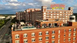 Rooftop party on a large brick hotel building with a rectangular pool and many people on the terrace; sign reads "South Side on Lamar" atop the structure with a bright blue sky and street below.