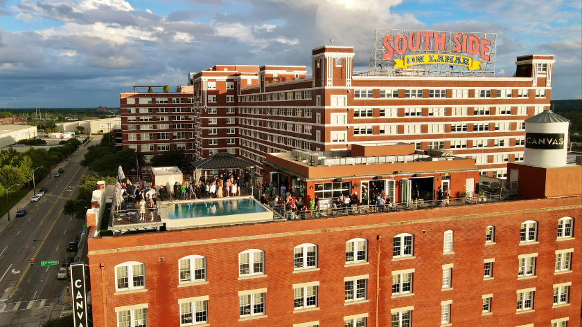 Rooftop party on a large brick hotel building with a rectangular pool and many people on the terrace; sign reads "South Side on Lamar" atop the structure with a bright blue sky and street below.