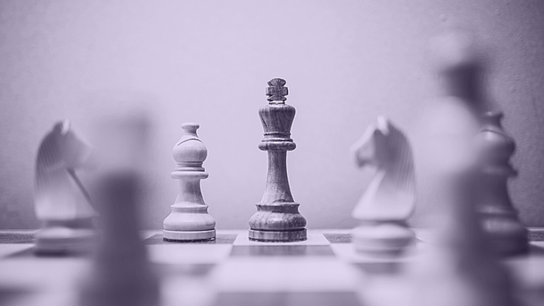 Wooden chess pieces on a chessboard with the king in sharp focus at center and blurred surrounding pieces in the background.