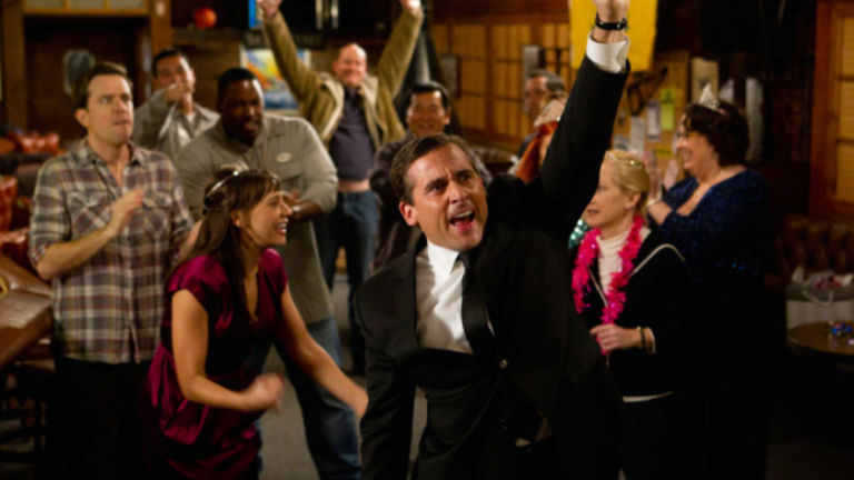 Man in a tuxedo cheers with a raised fist as colleagues cheer and dance around him at an indoor party district setting.