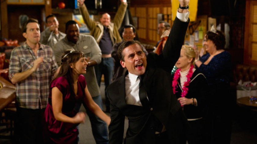 Man in a tuxedo cheers with a raised fist as colleagues cheer and dance around him at an indoor party district setting.