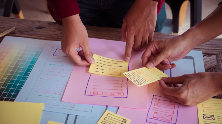 Group of hands arranging yellow sticky notes on a colorful UI storyboard is shown on a table a design board with sketches and swatches nearby.